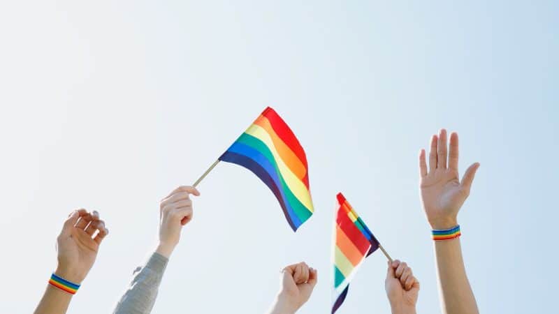 a blue sky with 5 peoples hands in the air waving a rainbow pride flag