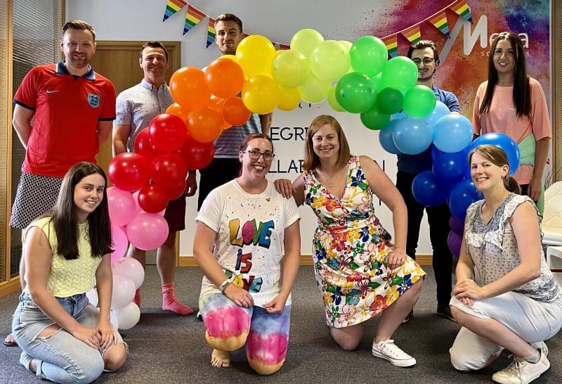 The Mexa team dressed in rainbow, standing underneath a rainbow balloon arch - celebrating Pride Month